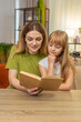 © Andrii Iemelianenko - Mother and daughter at home sit side by side alternate reading lines from child book smile and nod. Woman and girl at table keep pace help with words point page mark place enjoy calm time clear tone.