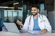 © Liubomir - Indian male doctor in white coat with stethoscope smiling and gesturing at laptop during a virtual video consultation, offering telemedicine advice and remote healthcare support