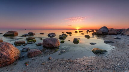Wall Mural - Rocks scatter along a serene beach as the sun sets on the horizon. The sky displays hues of orange and purple while water reflects the tranquil scene. Nature shows its beauty in this peaceful moment.