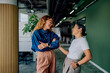 © we.bond.creations - Two diverse women colleagues laughing and talking in a modern office hallway, showcasing teamwork and professional friendship