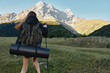 © SHOTPRIME STUDIO - A female hiker walks through a grassy meadow toward rugged mountains, carrying a rolled sleeping mat and a backpack, bathed in clear outdoor light.