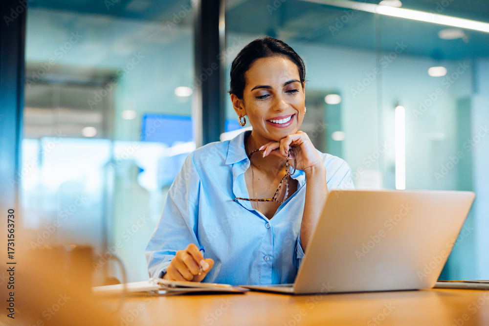 Smiling woman in office using laptop while holding glasses and working ...