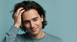 © mommy - Young man with long brown hair, hand on head, smiling, looking down and standing against a blue background