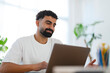 © Studio Marmellata - A young man with a beard works on his laptop in a bright, modern home office. He is focused and engaged with his computer, performing tasks or studying.