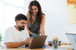 © Studio Marmellata - Two young colleagues are collaborating at a desk. The man shows something on his phone to the woman, who is smiling and looking at the screen. They are working together in a modern office.