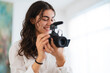 © Studio Marmellata - A smiling young woman with dark curly hair holds a professional camera with a microphone, adjusting settings or reviewing footage. She is focused and happy.