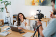 © Studio Marmellata - A smiling young woman sits at a desk with a laptop, holding a pencil, while a man films her with a camera. She is recording content for an online audience.