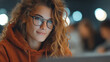 © SS Stock - A focused young woman with curly hair and glasses, working intently on her laptop in a dynamic, modern environment.
