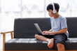 © Phimwilai - Young student wearing headphones using laptop on sofa at home for online education
