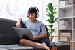 © Phimwilai - Asian boy wearing headphones using laptop on sofa at home