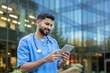 © Liubomir - Young male doctor smiling while looking at and using a digital tablet, wearing medical scrubs and a stethoscope in front of a modern hospital building