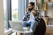 © sofiko14 - Man in his 30s speaking on phone while working remotely, using laptop and sitting at home