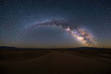 Wall Mural - A breathtaking panoramic scene of the Milky Way stretching over expansive desert sands at night, showcasing the galaxy's arching arm among the stars.