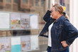 © Iryna - An elderly woman examines real estate advertisements displayed in a storefront window during the day
