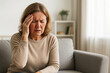 © Nataliia - Mature woman sitting on sofa with headache and pain expression, showing possible stroke or neurological disorder symptoms.