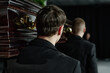 © AnnaStills - Caucasian young adult men carrying wooden coffin during funeral ceremony, walking in line with focus on back of heads, formal attire visible, somber indoor setting