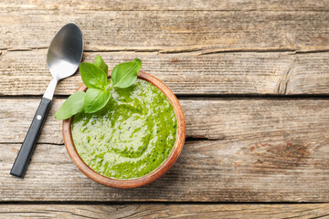 Wall Mural - Tasty green pesto in bowl and basil leaves on wooden table, top view. Space for text
