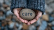 © Dinusha - Overhead photo of hands holding a smooth stone engraved with the word “care,” representing compassion, empathy, and mental health awareness.
