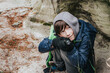 © Jolanta - A cheerful boy in a hoodie and gloves squats on the forest floor, holding a bright flashlight, near a mossy rock wall surrounded by sand and pine needles.