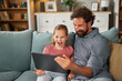 © Stockphotodirectors - A man and a young girl share a joyful moment as they watch a tablet together. The living room is filled with warm light, creating a cheerful atmosphere for their bonding time.