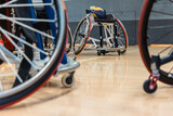 a close-up of wheelchair basketball players' chairs on an indoor court. A ball rests on one chair, with wheels and parts in focus against a blurred background of the gym floor and walls.