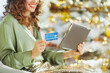 © Alliance - A happy woman with curly brown hair holds a credit card and tablet, engaged in online holiday shopping. Soft bokeh lights in the blurred background add to the warm, celebratory feel.
