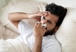 © Prostock-studio - Closeup Shot Of Sick Indian Man Blowing Runny Nose In Napkin While Lying In Bed At Home, Ill Young Eastern Male Touching Head, Feeling Unwell While resting In Bedroom, Having Seasonal Flu Or Fever