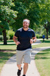 © AnnaStills - Caucasian young adult man jogging on park pathway, smiling and looking ahead, wearing headband and athletic clothing, trees and greenery in background, enjoying outdoor exercise