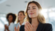 © StockHero - Young woman with long hair, wearing a dark blazer, is smiling and applauding in a bright conference room, surrounded by colleagues, celebrating a successful presentation or achievement