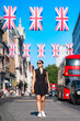 Young smiling Latina woman walking Oxford Street in London, England wearing a stylish black dress and sneakers, surrounded by Union Jack flags and red double-decker buses under a clear blue summer sky