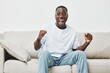 © SHOTPRIME STUDIO - Joyful young African man celebrating success in casual white shirt and denim jeans, expressing excitement with raised fists while sitting on a sofa