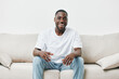 © SHOTPRIME STUDIO - Happy young Black man in casual outfit smiling on a beige sofa, conveying joy and relaxation in a cozy indoor setting