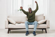 © SHOTPRIME STUDIO - Happy African American man celebrating success while sitting on a cozy sofa, wearing a casual green sweater and jeans in a bright, modern living room
