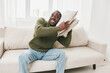 © SHOTPRIME STUDIO - Happy young man enjoying cozy time on the couch with pillows, wearing a green sweater and jeans, in a bright living room, emphasizing comfort and relaxation