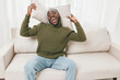 © SHOTPRIME STUDIO - Happy African American man in green sweater smiling with pillows on white couch indoors, showcasing joy and comfort in modern lifestyle