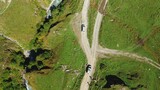 A top-down aerial view of two cars of travelers driving along a dirt road one after the other.