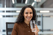 © Liubomir - Young professional woman with curly hair smiling at the camera while drinking a glass of pure water in a modern office, staying hydrated and refreshed during her workday