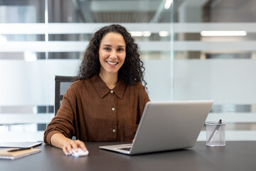 Young businesswoman with curly hair smiling at camera, using a laptop and mouse while sitting at a dark desk in a contemporary corporate office environment