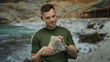 © Krakenimages.com - Young man with tattoos holding us dollars by the seaside at the beach, reflecting wealth and leisure in a coastal outdoor setting under the sun.