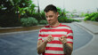 © Krakenimages.com - Young man with tattoos wearing striped shirt stands outdoors with a playful expression on a city street showing gestures surrounded by urban greenery.
