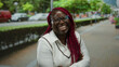 © Krakenimages.com - Smiling african american woman with braided hair and glasses standing on a city street with arms crossed, surrounded by urban greenery and passing cars on a sunny day.
