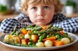 © Alla - Child with blonde hair in checkered shirt sits at wooden table, staring at colorful plate of vegetables, showing refusal to eat healthy food, expressing disinterest in meal