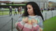 Young hispanic woman in colorful sweater expresses surprise while pointing at herself against the backdrop of pisa tower, outdoor tourist setting with blurred crowd in italy.