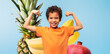 © Prostock-studio - Young boy in an orange t-shirt proudly shows off his muscles with a big smile. The soft blue background highlights his confidence, complemented by colorful fruit elements like pineapples and bananas.