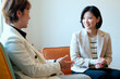© pressmaster - Two Asian women sitting and having conversation, one middle aged woman gesturing while talking, young adult woman listening and smiling, both wearing business attire in office setting