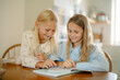 © m-art - Two girls share a joyful moment while reading a book together at a warm, inviting kitchen table. They are engaged and smiling, enjoying each other's company.