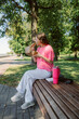 © m-art - A young woman sits on a wooden bench in a green park, enjoying her meal from a takeout box. The sun shines brightly as she hugs her drink and takes a bite, surrounded by nature.