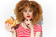 © VICHIZH - Woman with curly hair enjoying a burger, making a playful gesture with her finger to her lips, wearing a striped shirt, isolated on a white background