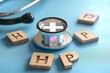 © Olya - Close-up of a silver stethoscope with a medical cross symbol reflected on the diaphragm, surrounded by wooden letter tiles forming the abbreviation with some letters scattered on a blue surface.