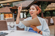 © EdNurg - Woman holding menu, sitting casually at outdoor restaurant table, raising hand to call for waiter